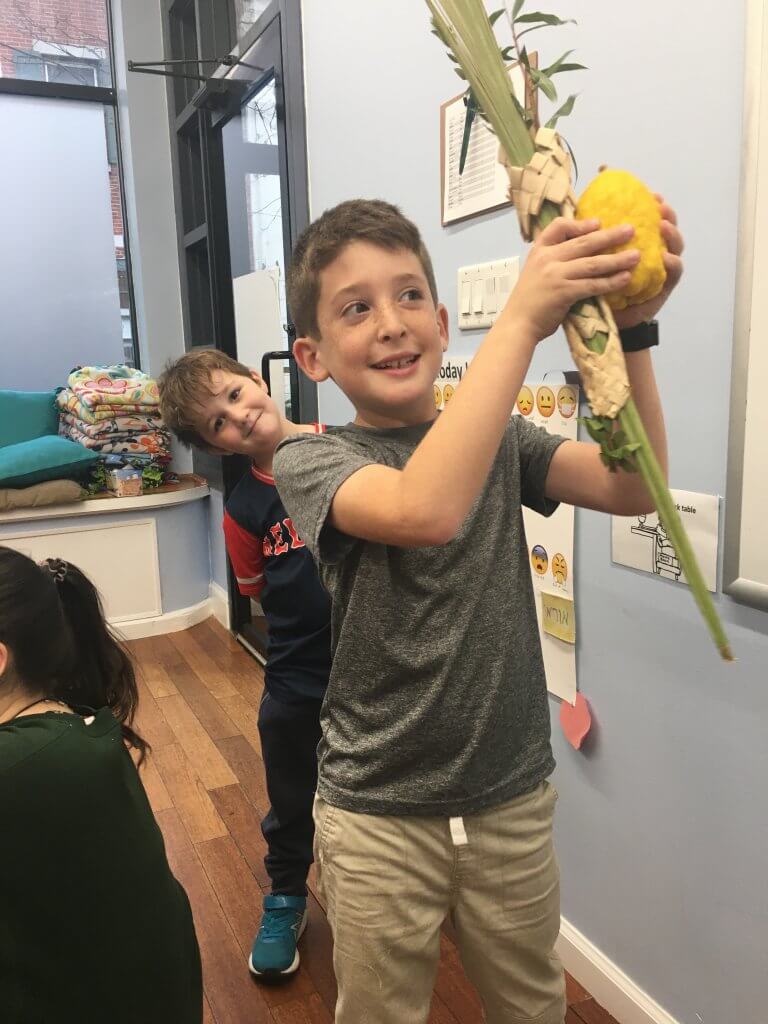 A 2nd grader shakes lulav while a 1st grade friend photobombs.