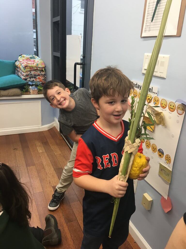 A 1st grader shakes lulav while a 2nd grade friend photobombs.